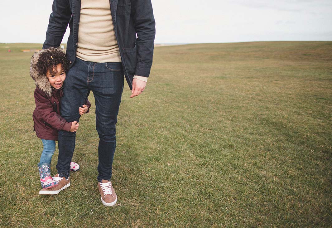 Young girl holding her fathers leg, standing in an open field.