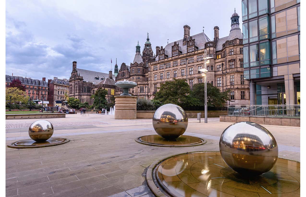 The Millenium Square in Sheffield, and the large golden globe water fountains, against a brack drop of a large stately building.
