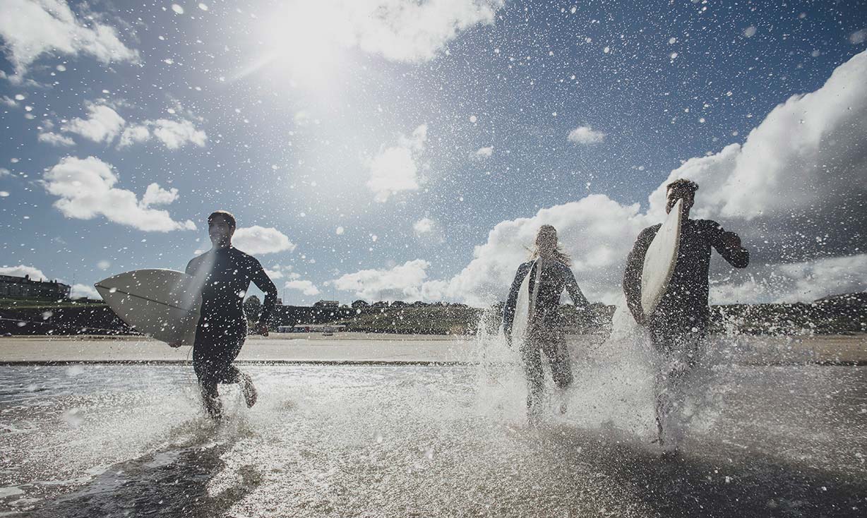 Three surfers carrying surfboards running into the sea, splashing everywhere.