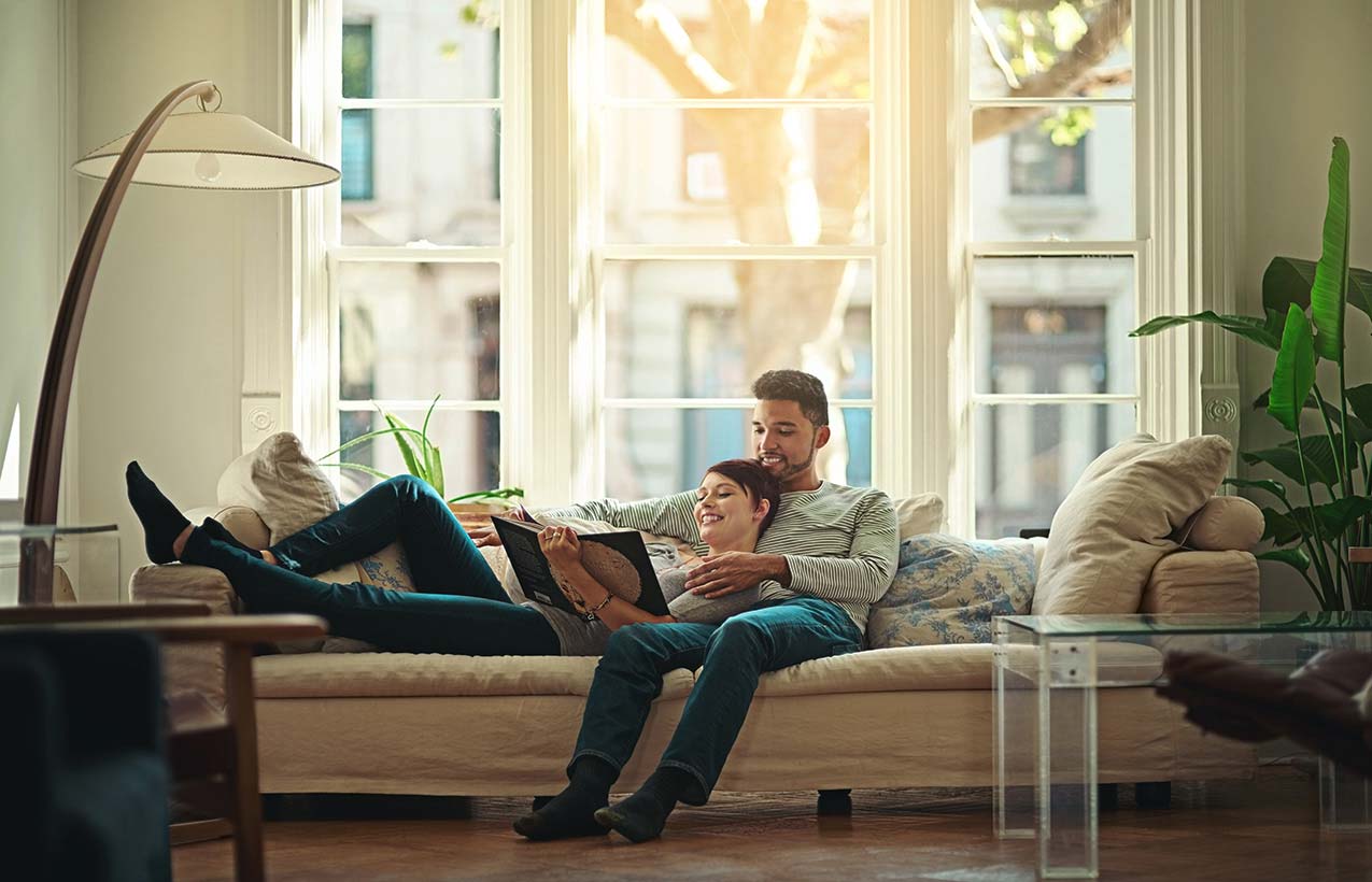 Couple enjoying time together lying on the sofa. The woman is reading a book whilst reclining on her partner. Sunlight pours through the window behind them.