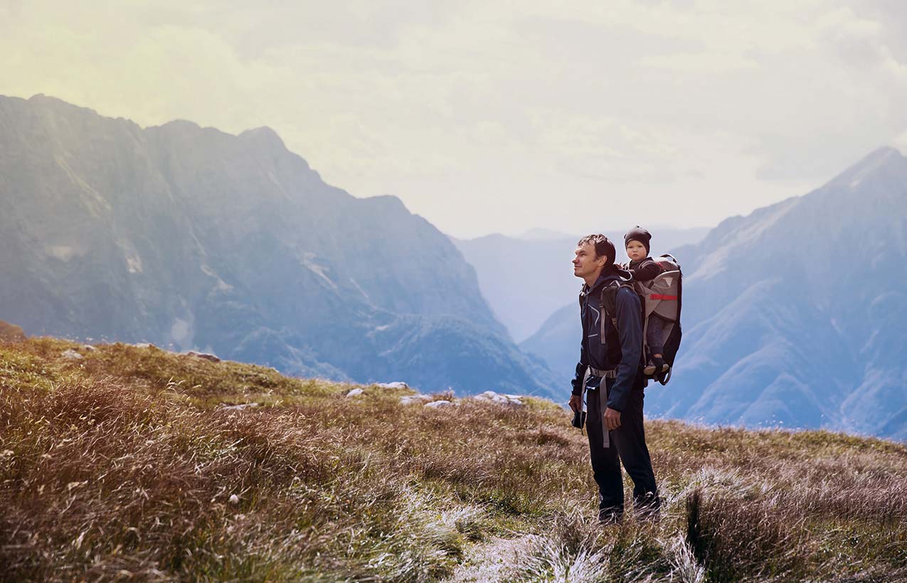 Man and young child travelling on a mountain. The young child is carried on the man's back and they're looking at the camera.