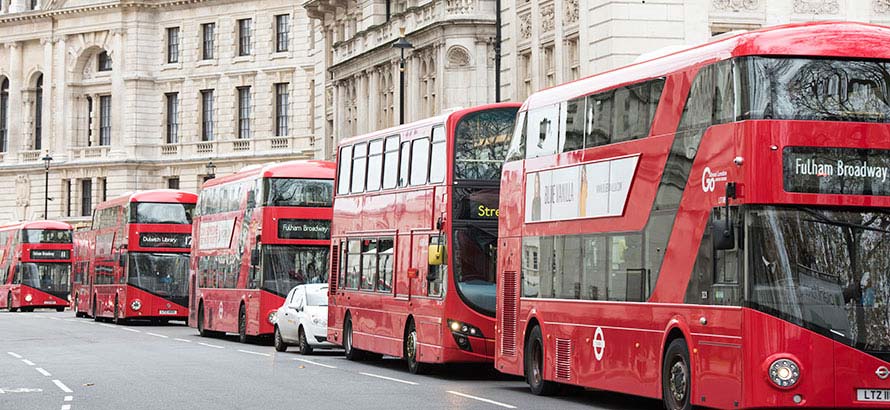 A side view of a line of red London buses along a road, with sandstone townhouses in the background.
