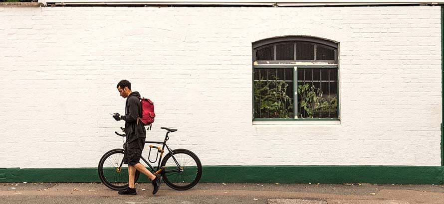 Young man pushing his bicycle along a road in Hackney, with white washed walls of in the background.