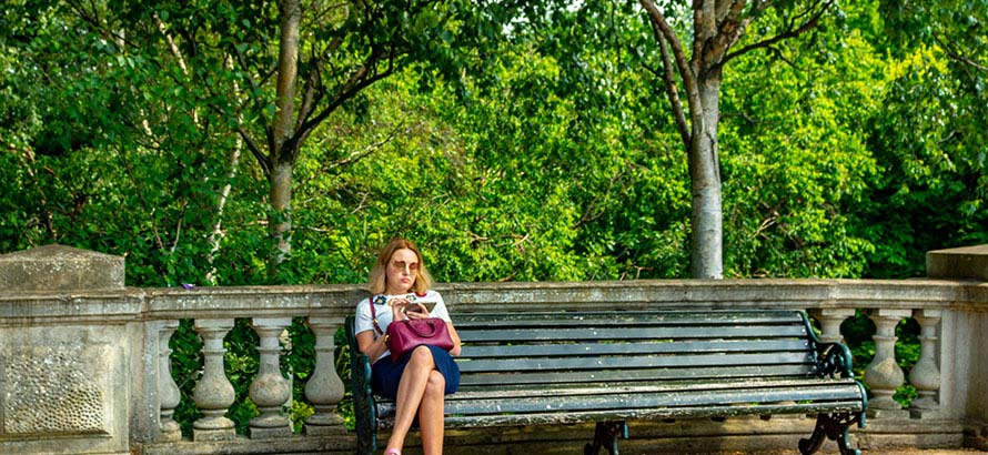 Woman sitting on a wooden park, reading, against a backdrop of leafy trees.