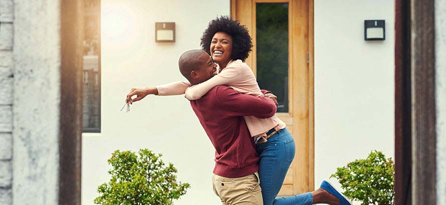 Couple happily embracing, with the man lifting the woman off the floor, outside of a front door to a home.