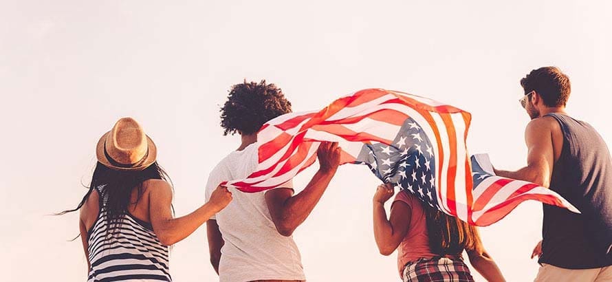 Four friends walking away from the camera, holding out a large American flag behind them.